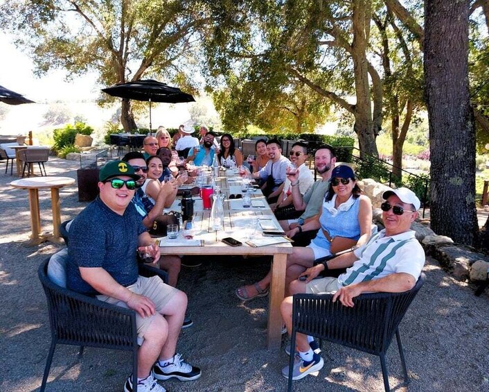 A group of people seated at an outdoor table with drinks, surrounded by trees and outdoor furniture. 5