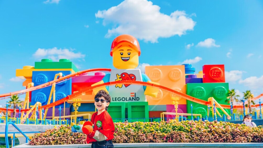 A child in a red shirt holding a toy in front of a LEGO-themed building with colorful blocks and a roller coaster. 4