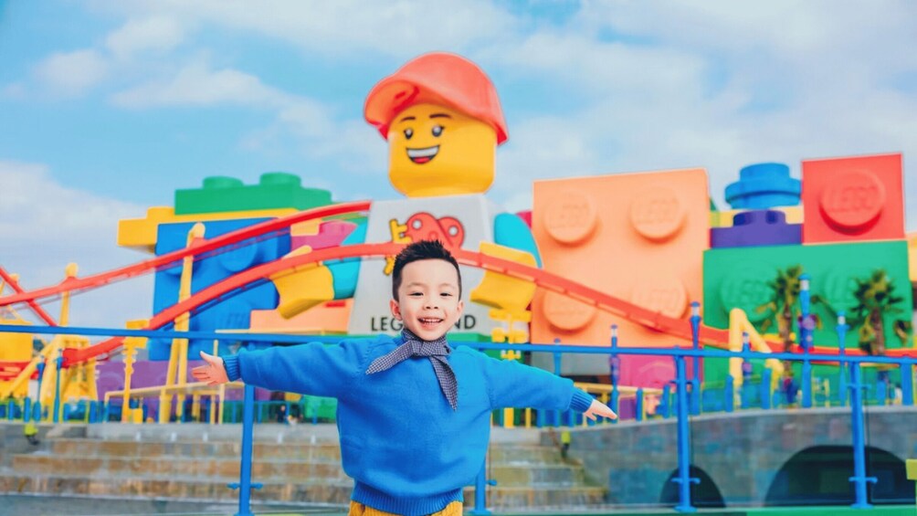 A child in a blue sweater with arms outstretched in front of a LEGO-themed play area with colorful structures and a bridge. 1