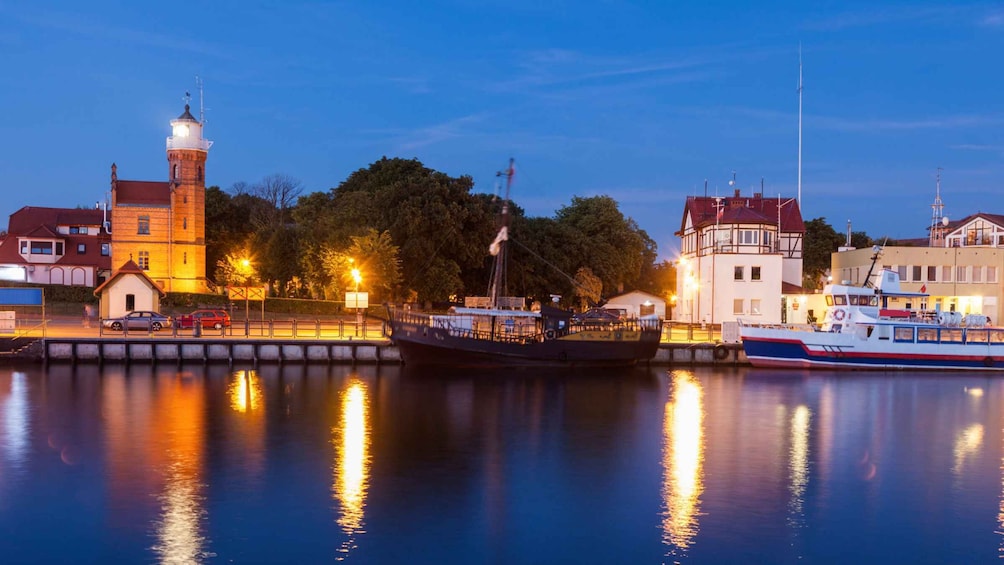 A waterfront with a historic tower, a modern building, and boats docked at a marina. 1