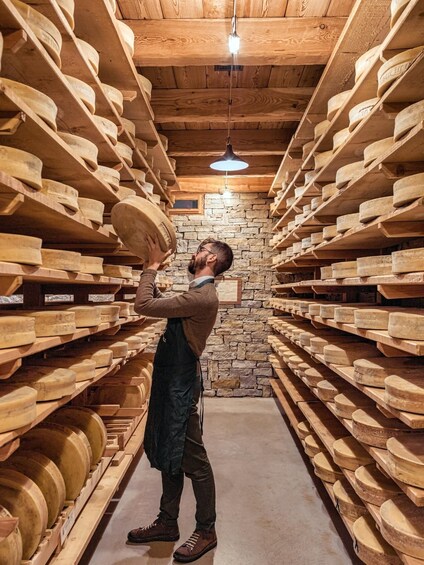 A person inspecting cheese wheels in a wooden cellar with stone walls and wooden shelves. 1
