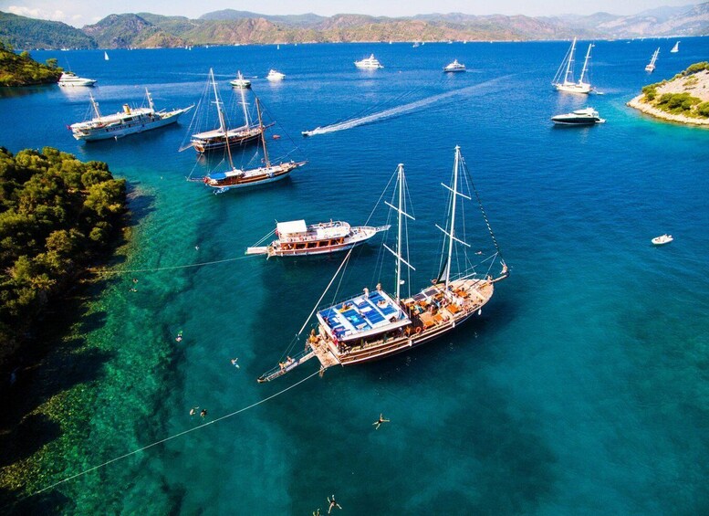 A coastal area with several sailboats anchored in clear blue waters, surrounded by lush greenery and distant hills. 1