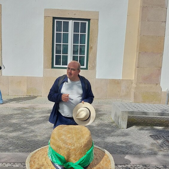 A man standing in front of a building with a window, holding a hat. 5