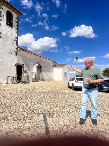 A man standing in front of a historic building with a cobblestone courtyard, a white car, and a clear blue sky. 2