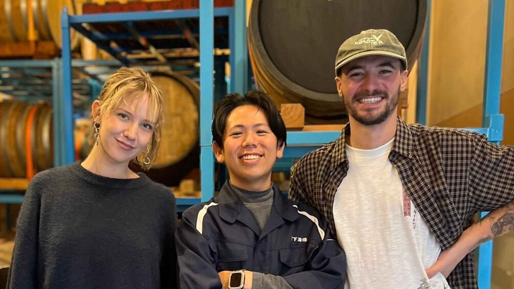 Three people standing in a room with large barrels in the background. 1