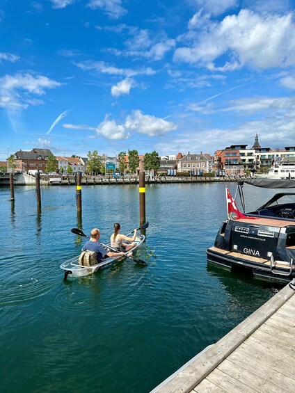 A calm river with a boat carrying two people, a dock, and buildings in the background. 1