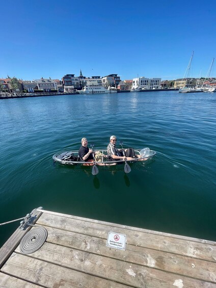 Two people rowing a small boat on a calm river with buildings in the background. 2