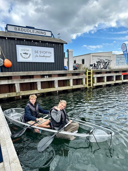 Two people in a small boat at a dock with a sign that reads 'BÄDUDLEJNING' and 'SE SYDFYN fra Svenborgsund'. 3