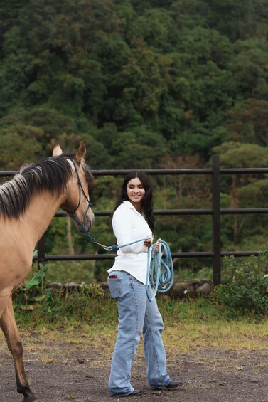 A person leading a horse with a blue rope, standing in a fenced area with greenery in the background. 2