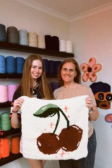 Two people holding a knitted item with a cherry design. In the background, there are shelves with various colored yarns and a wall with a flower and a cartoon character. 2