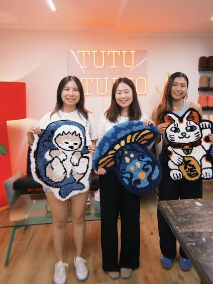 Three people holding hand-knitted cat-shaped blankets in front of a store named 'TUTU STUDIO'. 1