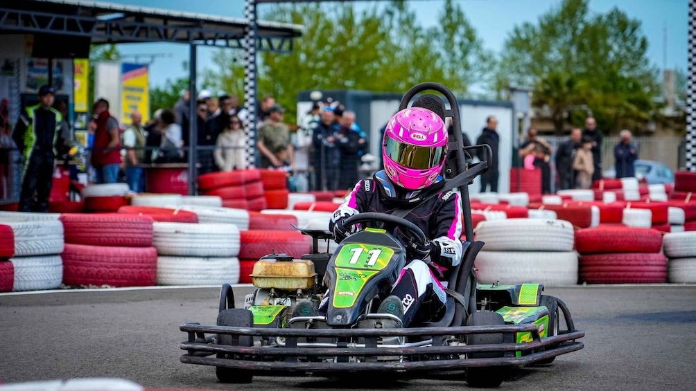A go-kart racer in a pink helmet and black suit driving on a track with spectators in the background. 1