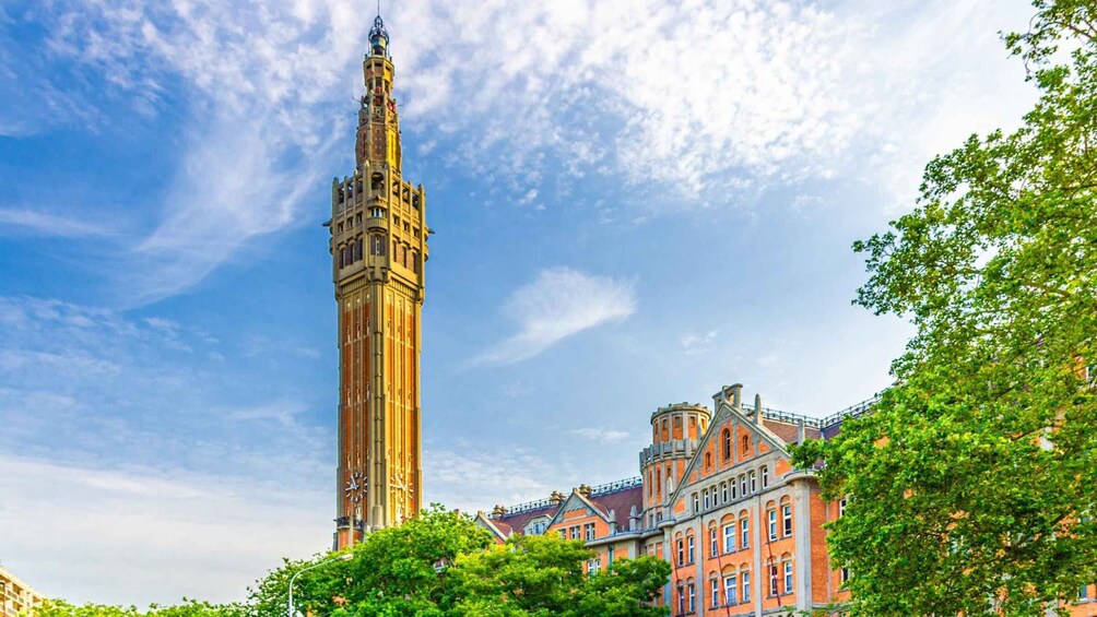A tall clock tower with a pointed spire, surrounded by lush green trees and historic buildings with red brick facades. 1