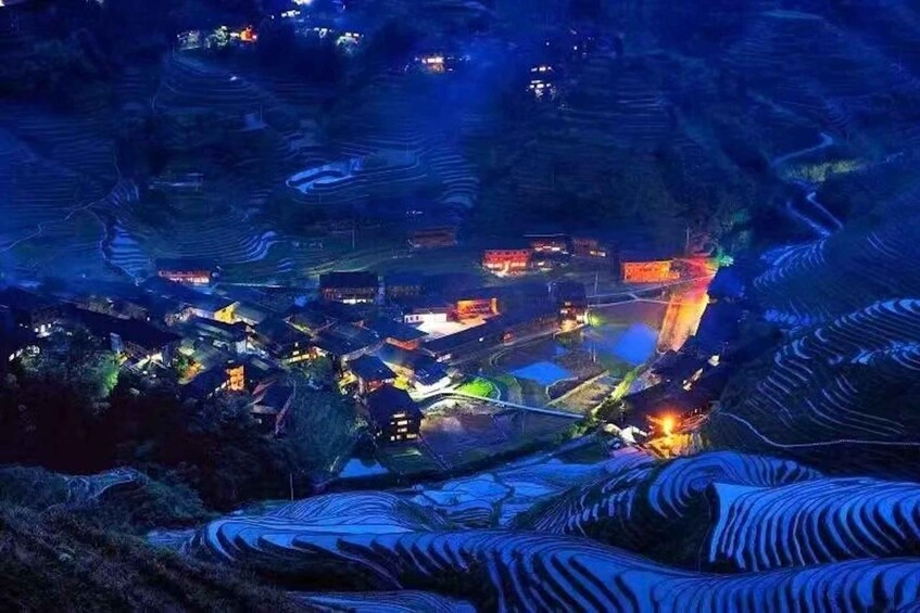 A nighttime aerial view of a village with terraced fields and illuminated buildings. 3