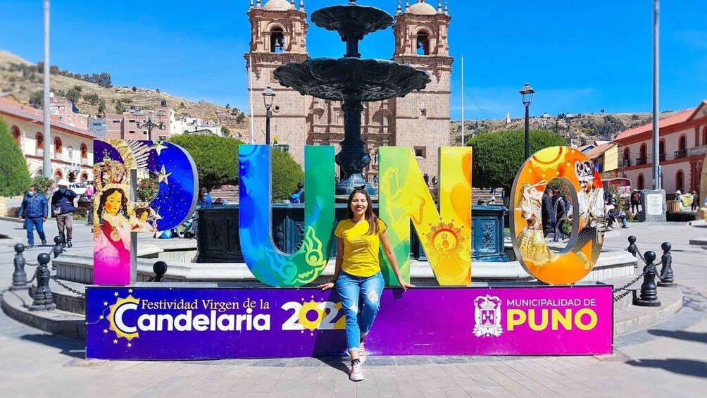 A woman standing in front of a colorful sign with the letters 'PUNO' and 'Festividad Virgen de la Candelaria 2021' in a town square with a fountain and a church in the background. 3