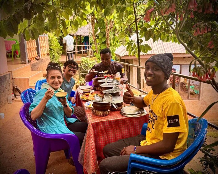 A group of people sitting at an outdoor table with plates of food, surrounded by greenery and a thatched-roof structure. 2