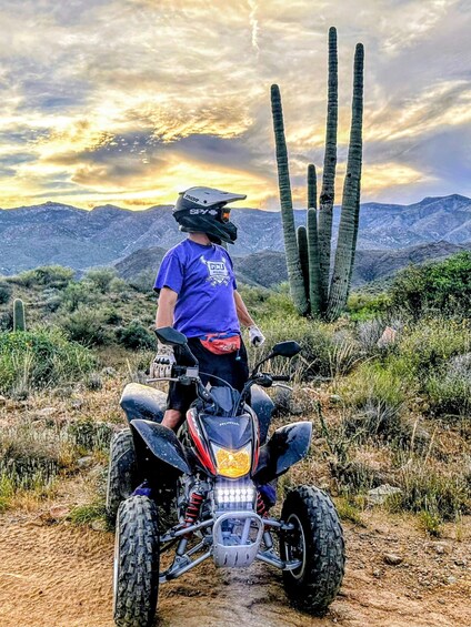 A person riding a quad bike on a dirt trail with cacti and mountains in the background. 3