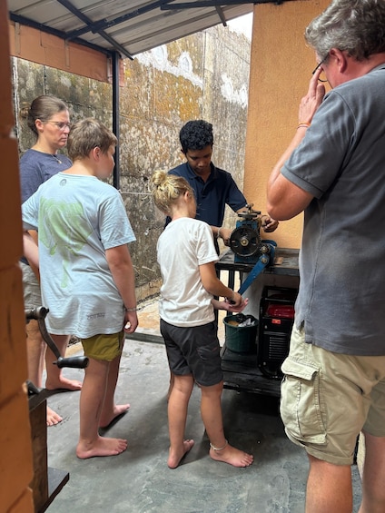 A group of people around a mechanical device, possibly a generator, in an outdoor setting with a concrete floor and a metal roof. 2