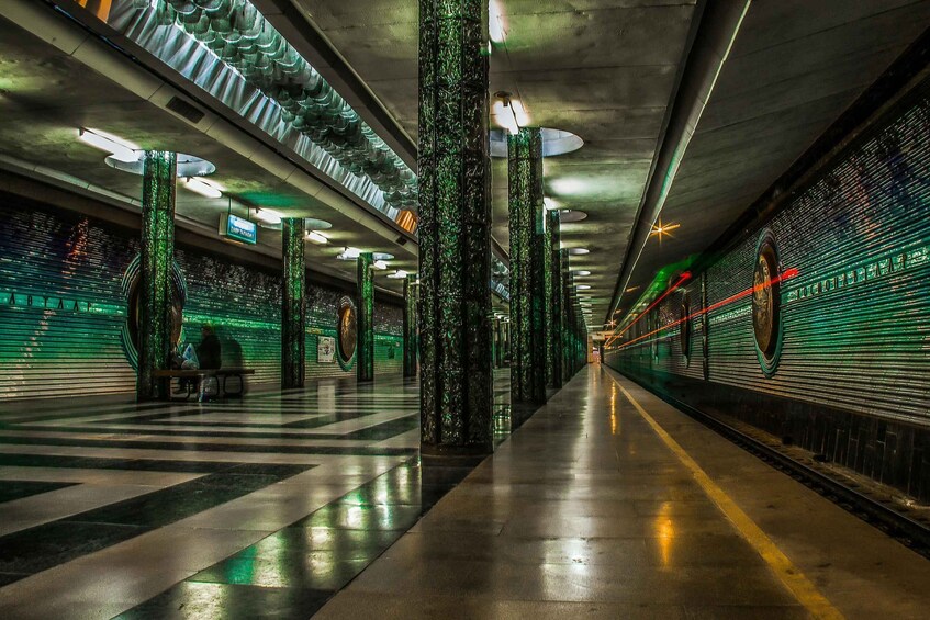 A subway station with green lighting, a train on the platform, and a staircase. 3