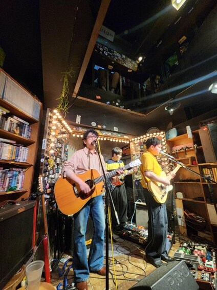 A musician playing an acoustic guitar, another playing an electric guitar, and a third playing a bass guitar in a room with shelves of books and various items. 1