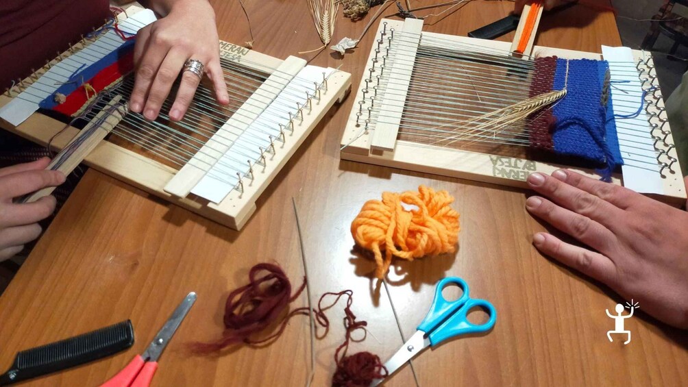 Two individuals weaving on wooden looms with yarn, scissors, and a comb on a wooden table. 2