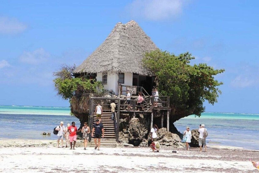 A thatched-roof structure on a beach with clear blue water and a few people around it. 5