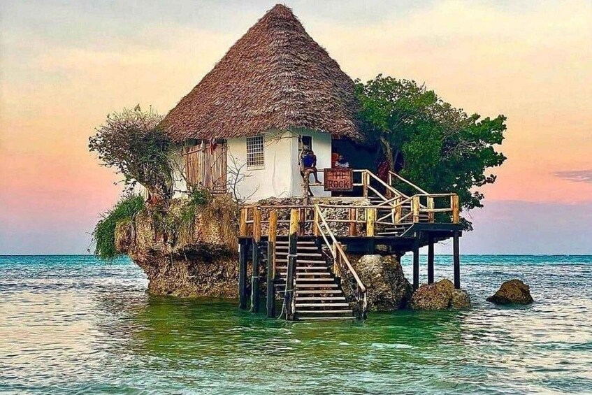 A thatched-roof house on stilts over water with a wooden staircase leading up to it. 1
