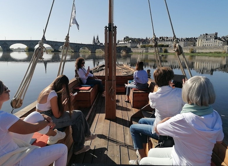 A group of people on a wooden boat with ropes, a bridge, and buildings in the background. 1