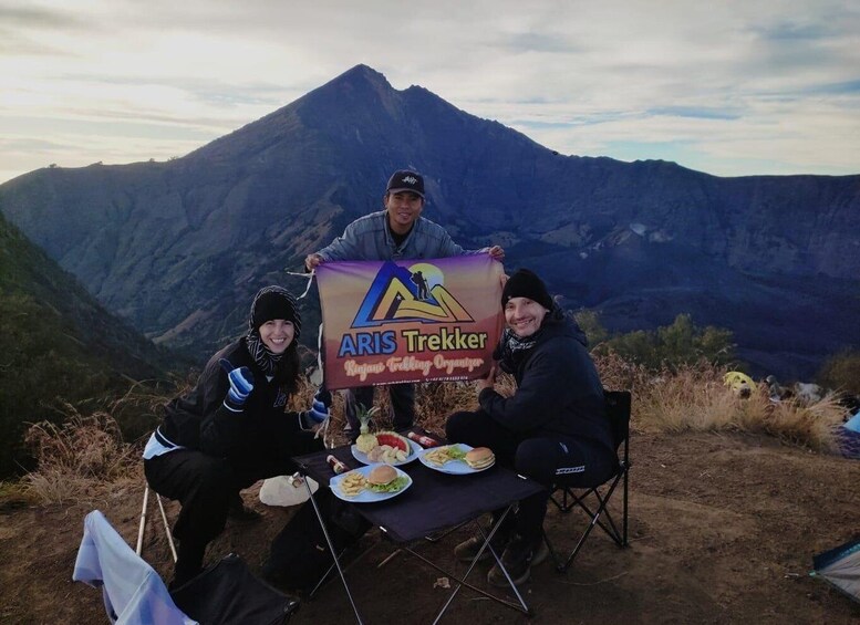 Three people sitting at a table with food, holding a banner with a mountain in the background. 1