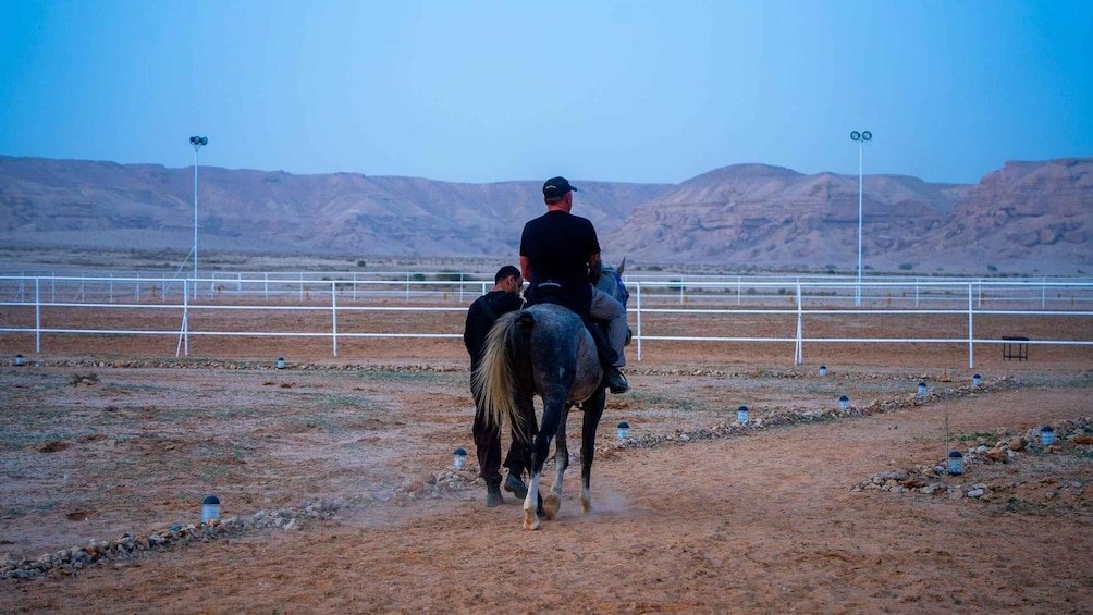 Two riders on horses in a fenced arena with mountains in the background. 4