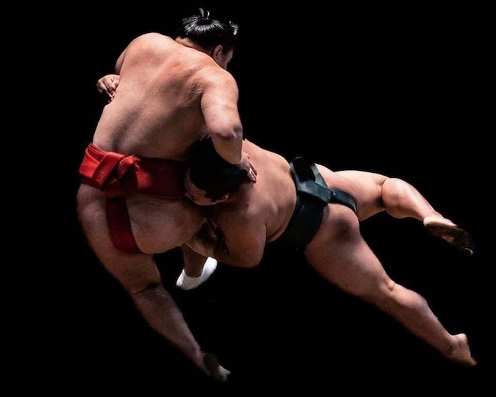 Two sumo wrestlers in a match, one in red and the other in black, in mid-air. 1