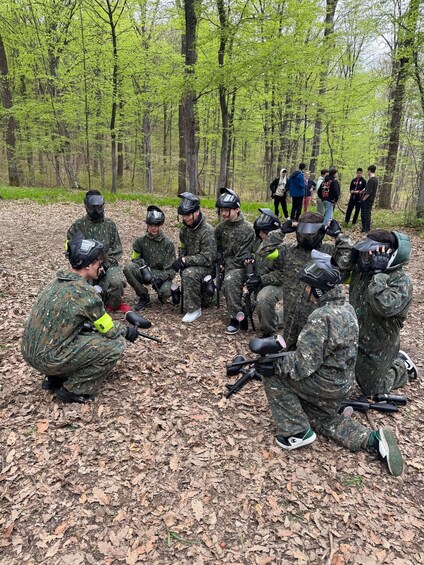 A group of people in camouflage uniforms and paintball gear sitting in a circle on a forest floor. 3