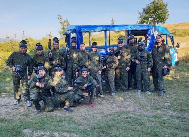 A group of people in military camouflage uniforms posing with paintball guns in an outdoor setting with a blue vehicle in the background. 2