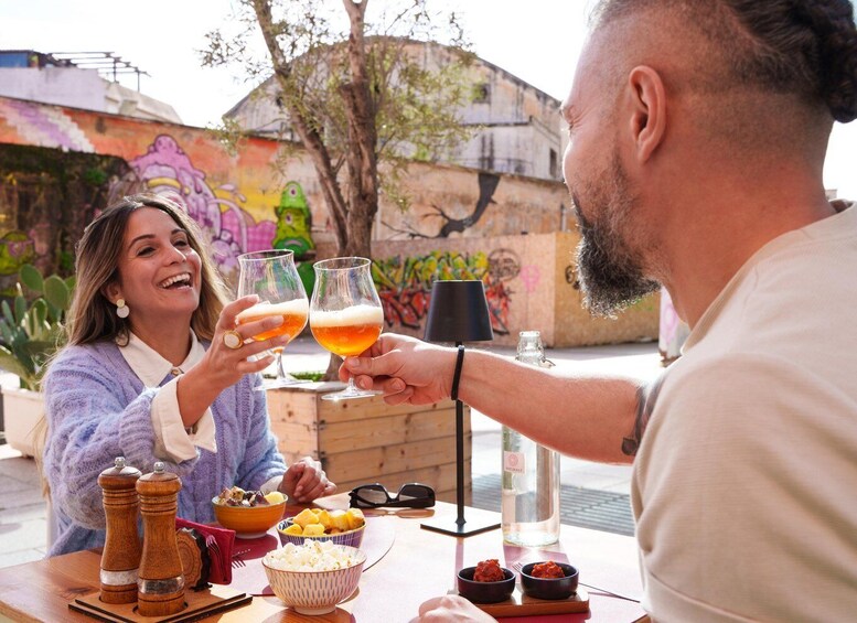 Two people toasting with glasses of beer, a table with food and condiments, and a graffiti-covered wall in the background. 1