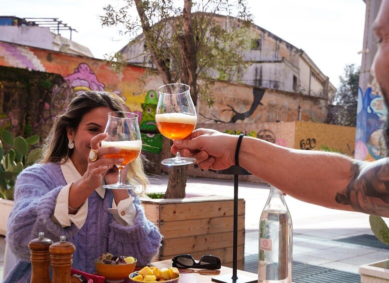 Two people toasting with drinks, a table with food and a bottle, and a colorful mural in the background. 5