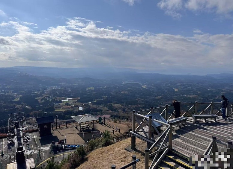 A panoramic view from a high vantage point overlooking a valley with a wooden observation deck, a small shelter, and distant hills. 2