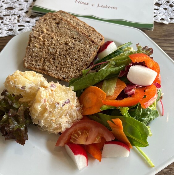 A plate with a slice of whole grain bread, a serving of potato salad, and a mixed vegetable salad with radishes and carrots. 2