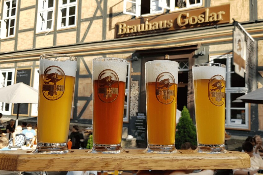 Four glasses of beer with logos on a wooden tray, placed in front of a building with a sign that reads 'Brauhaus Goslar'. 3