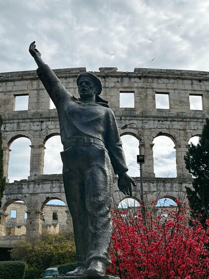 A statue of a man with a raised arm, a stone building with arches in the background, and red flowering bushes. 4