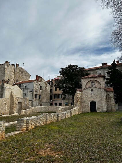 Ancient stone buildings with a 'For Sale' sign, a grassy area, and a cloudy sky. 2