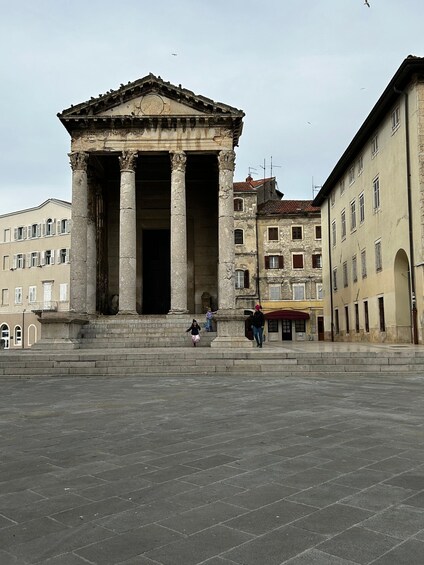 A classical building with columns and a pediment, surrounded by a paved square and adjacent buildings. 5