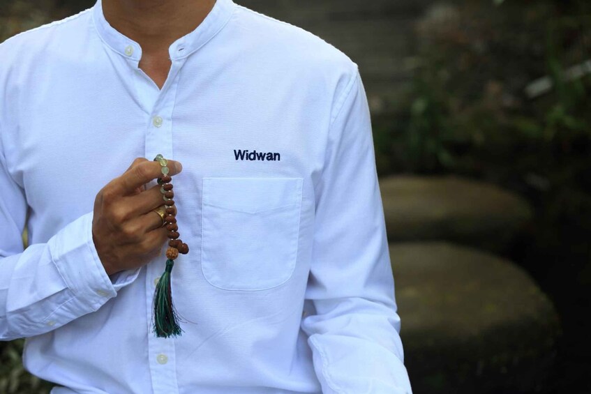 A person wearing a white shirt with the word 'Widwan' on the chest pocket, holding a string of prayer beads. 3