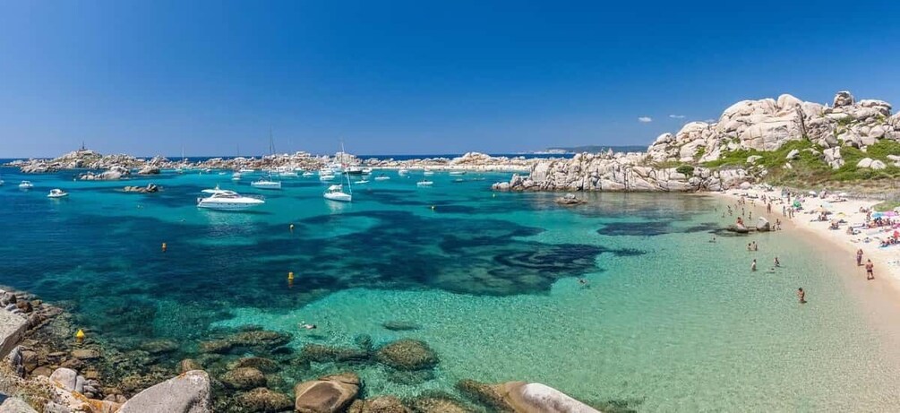 A coastal area with clear turquoise waters, rocky formations, and a sandy beach with people. 2