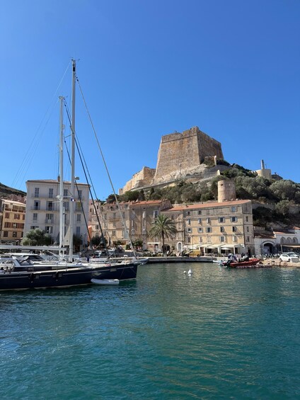 A coastal town with a prominent fortress on a hill, a sailboat docked at the marina, and buildings along the waterfront. 1