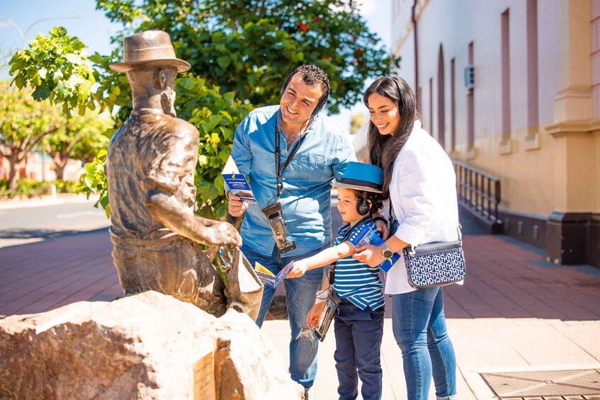 A family interacting with a bronze statue of a man in a hat, holding a book, and a child wearing a striped shirt and hat. 1