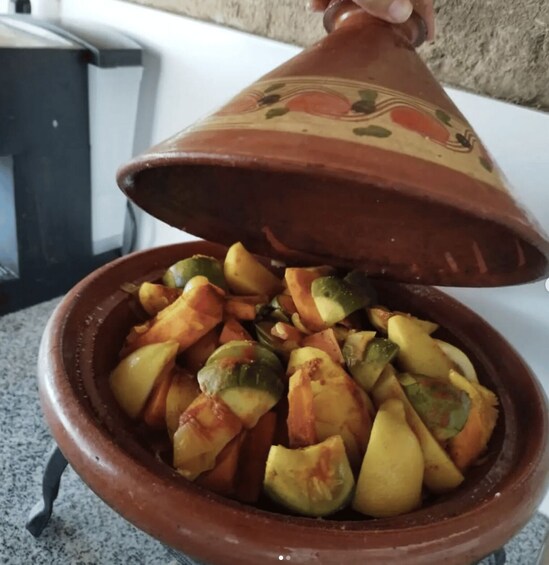 A hand holding a traditional Moroccan tagine pot with a lid, filled with sliced fruits. 1