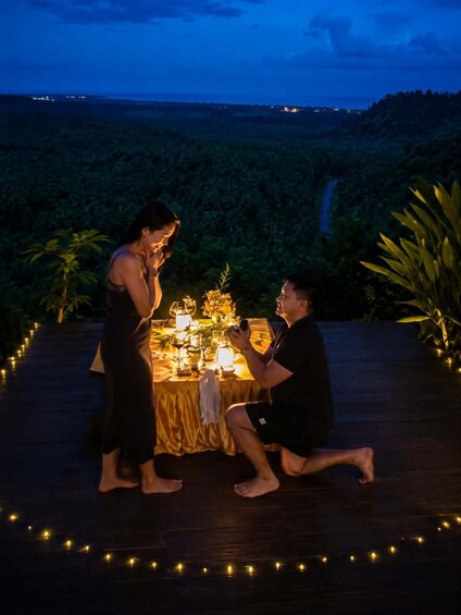 A couple on a wooden deck with a table set for a romantic dinner, surrounded by greenery and string lights. 5