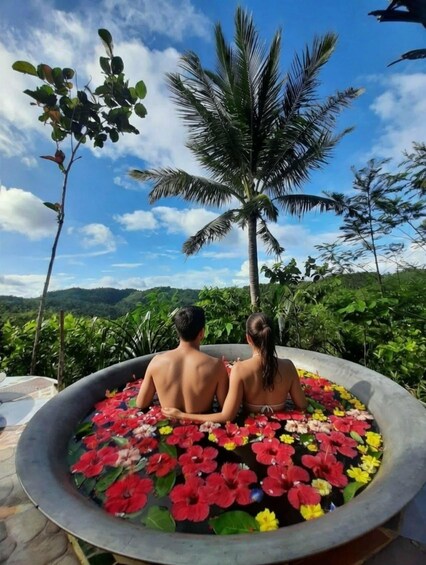 Two people sitting in a hot tub filled with flowers, surrounded by lush greenery and a palm tree. 3