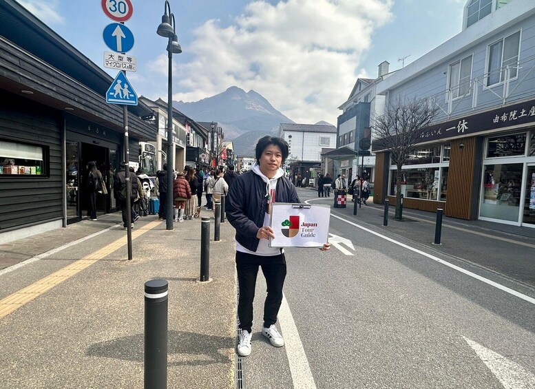A person holding a Japan Tour Guide sign on a street with shops and a mountain in the background. 5