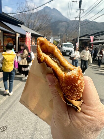 A hand holding a fried dough food item with a visible filling, set against a street scene with people and mountains in the background. 3